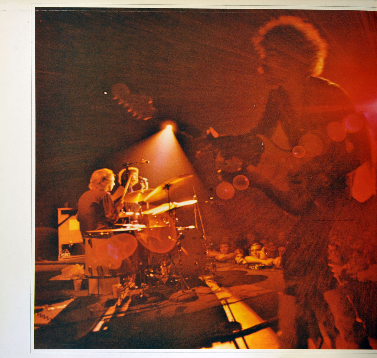 Jimi Hendrix performing live, silhouetted against deep red and orange stage lighting. His blurred figure and glowing afro add to the ethereal, electrifying atmosphere of the performance. In the background, the drummer, illuminated by a spotlight, is captured mid-motion, with the audience watching in awe from the edge of the stage. The lens flare and dramatic lighting enhance the raw energy and intensity of the moment, immersing the viewer in the concert’s fiery ambiance.